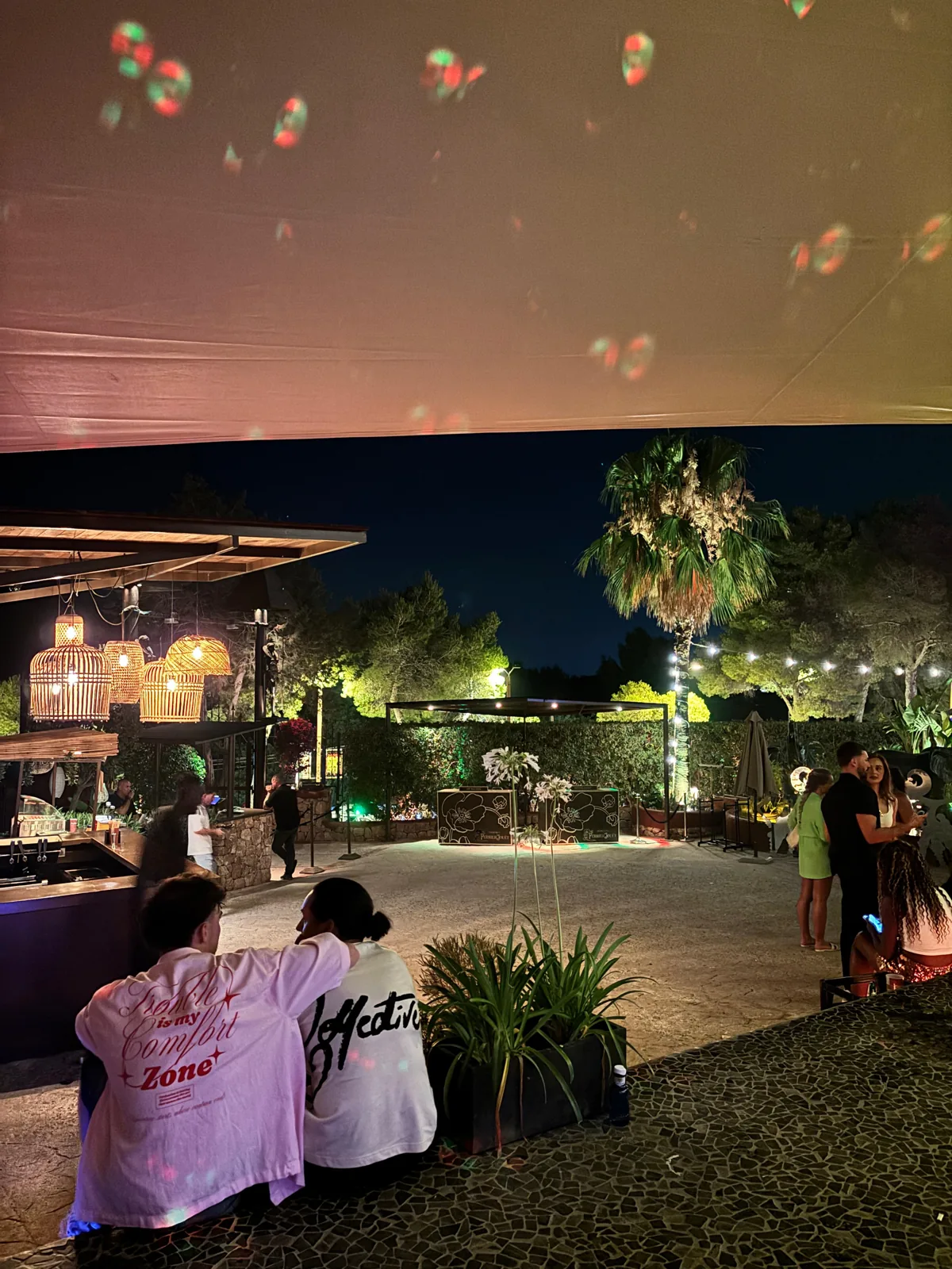 Gäste im Open-Air-Bereich der Cova Santa mit farbigen Lichtprojektionen und Palmen bei Nacht