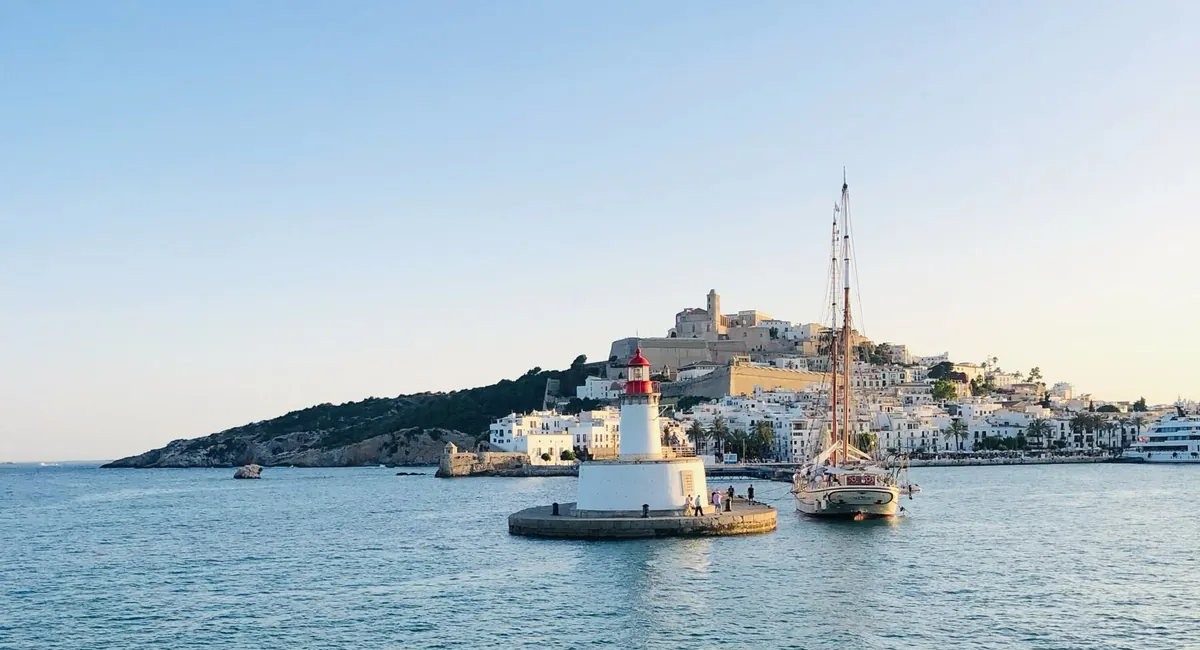 Harbor lighthouse at the entrance to the port of Ibiza Town with Dalt Vila and a sailing ship in the evening light