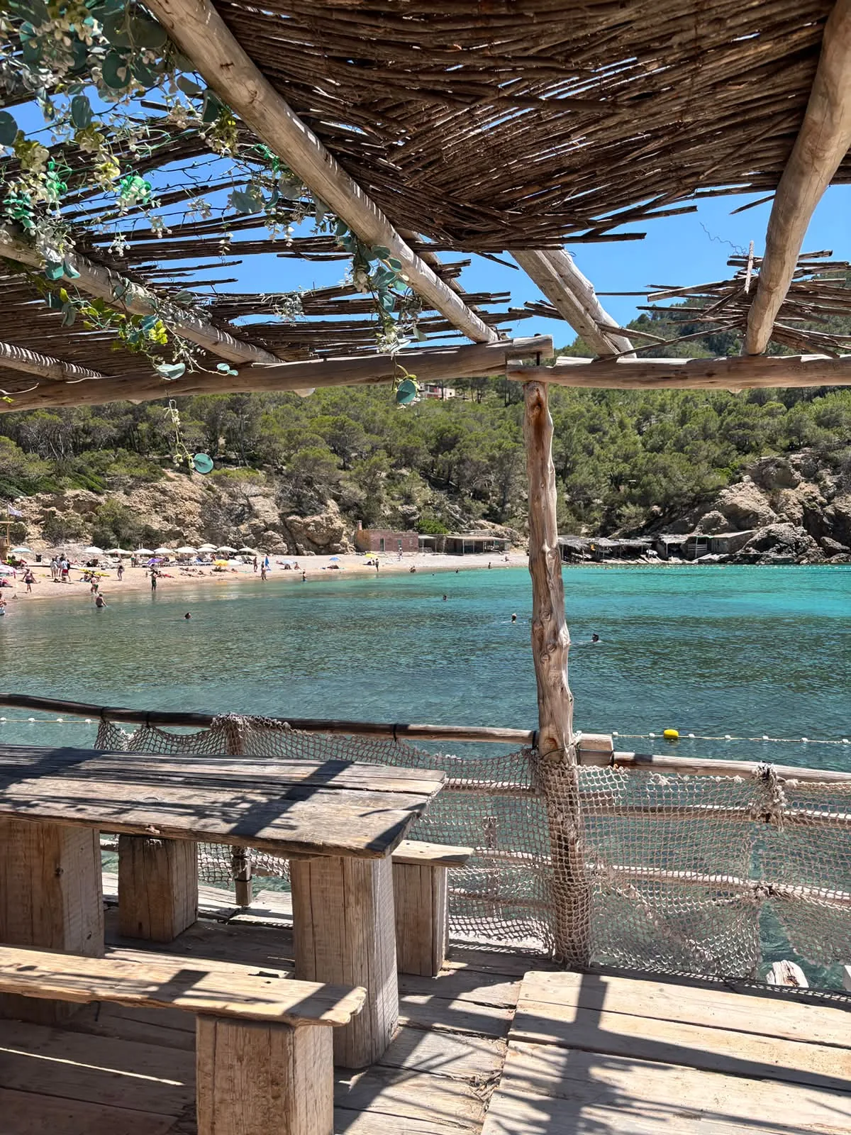Blick von einem Strandlokal mit Holzdach über die Bucht der Cala Benirràs auf Strand und türkises Meer