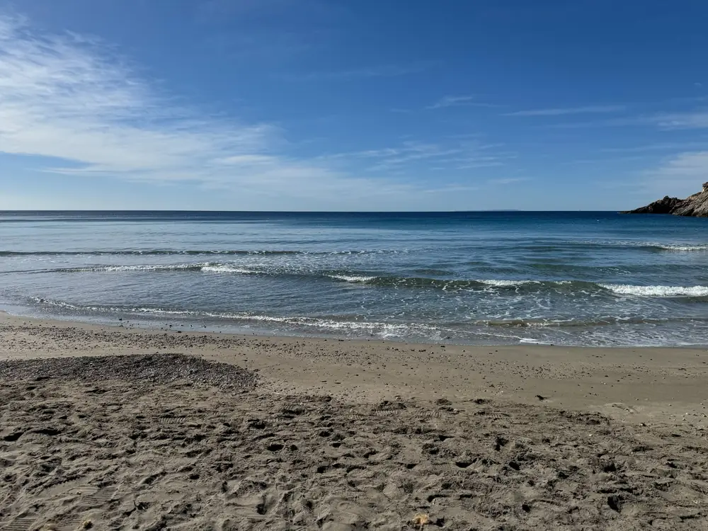 Dunkler Sand der Cala Boix mit ruhiger Brandung und tiefblauem Meer