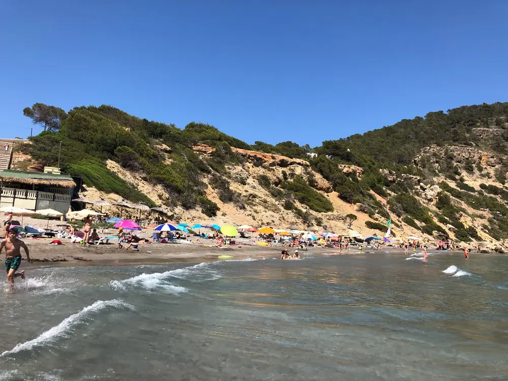 Strand der Cala Boix mit bunten Sonnenschirmen, Strandbar und bewaldeten Klippen