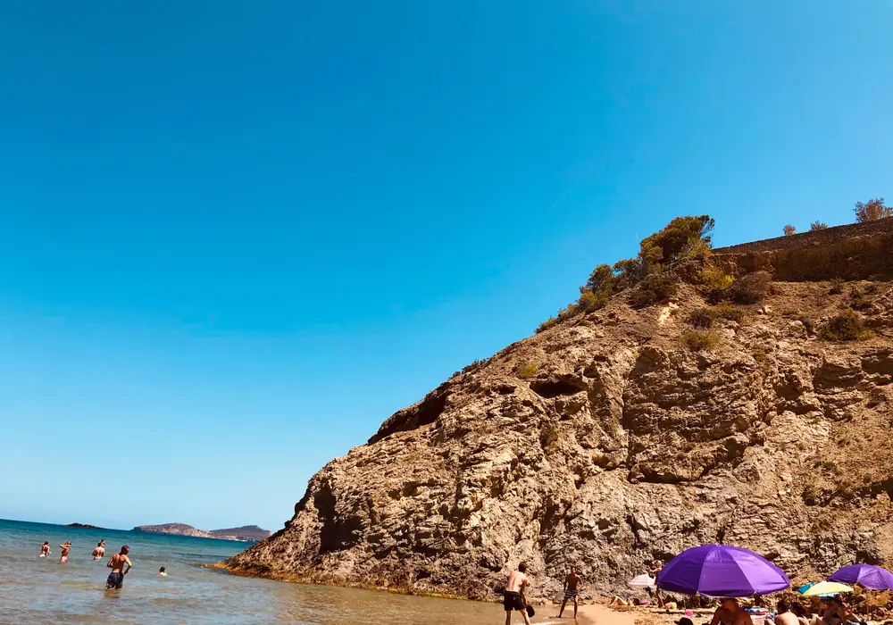 Badende und Sonnenschirme am Strand der Cala Boix unter dem markanten Felsmassiv