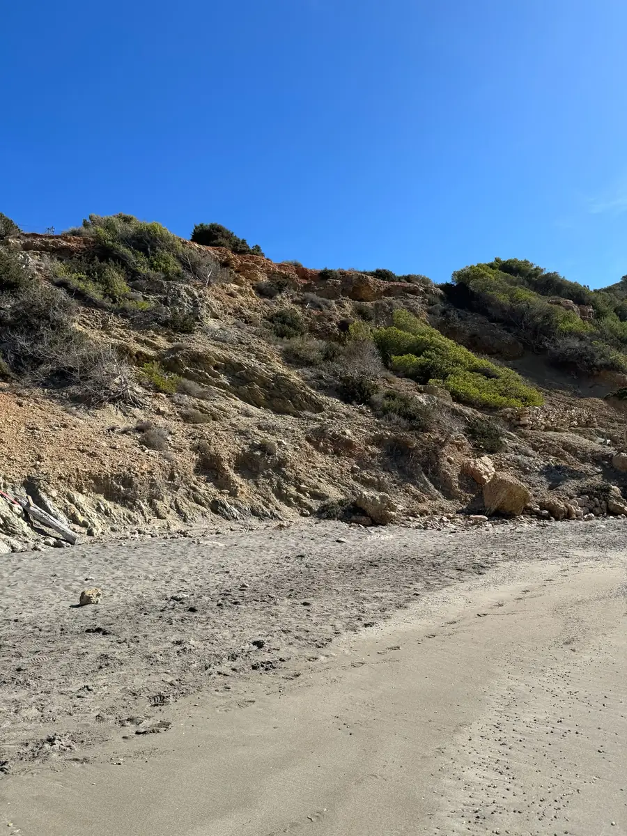 Dunkler, fast schwarzer Sand der Cala Boix vor der rot-ockerfarbenen Steilküste