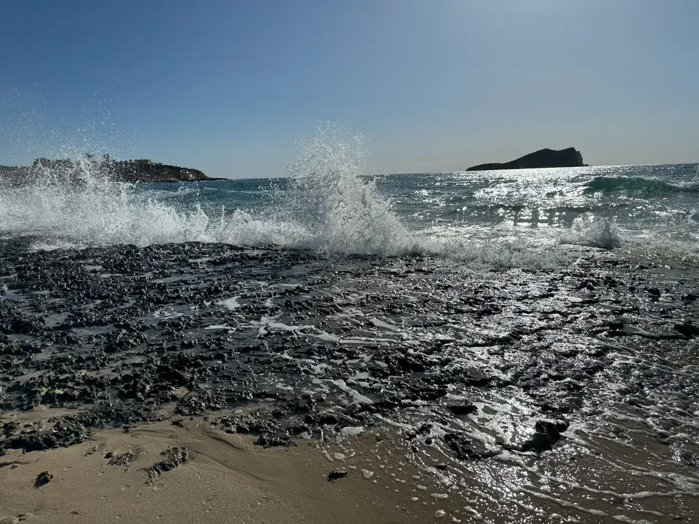 Brandung über dunklen Felsen an der Cala Comte mit vorgelagerter Insel