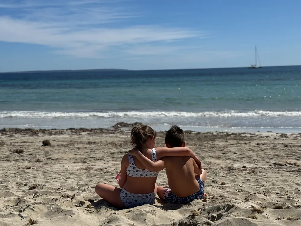 Two children sit arm in arm on the sandy beach of Ses Salines and look out at the turquoise-blue sea with a sailboat on the horizon