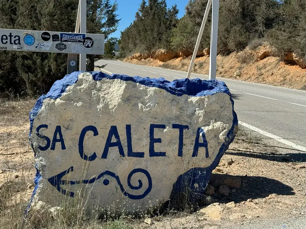 Bloc de roche peint en bleu et blanc avec l’inscription 'Sa Caleta' et une flèche comme panneau indicateur au bord de la route